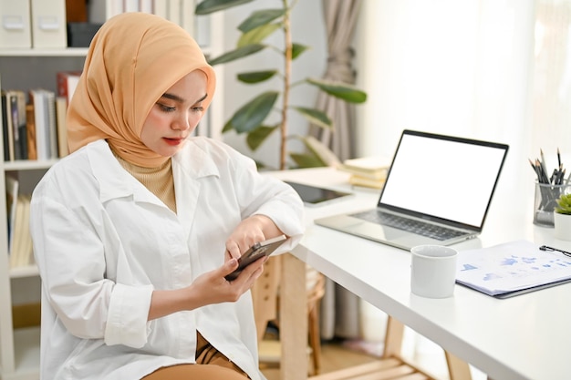 Mulher de negócios muçulmana asiática profissional usando seu telefone em sua mesa no escritório moderno