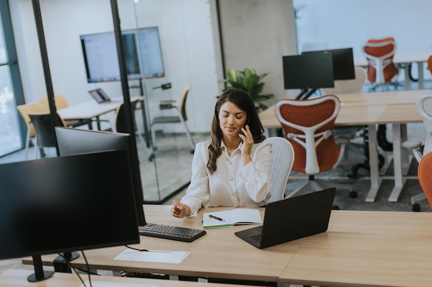 Mulher de negócios muito jovem usando telefone celular enquanto trabalhava no laptop no escritório moderno