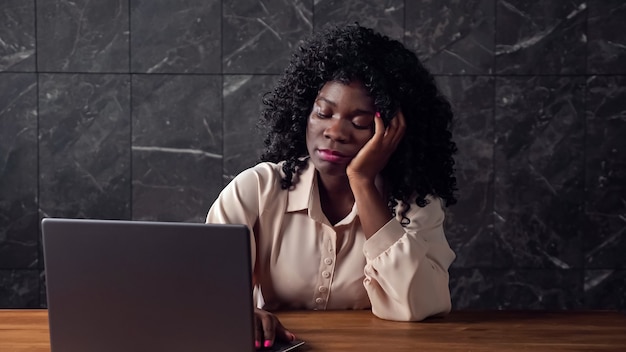 Mulher de negócios negra exausta com cabelos cacheados em um laptop cinza preguiçosamente e boceja sentada à mesa de madeira marrom no escritório tarde da noite
