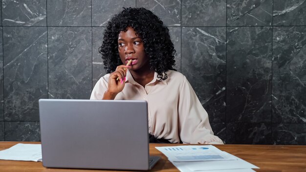 Mulher de negócios negra preocupada e pensativa com cabelo encaracolado olha em volta sentada no laptop cinza com folhas de papel e uma caneta roendo no escritório