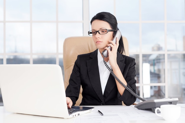 Mulher de negócios no trabalho. Mulher de negócios jovem confiante falando ao telefone e usando o computador enquanto está sentada no local de trabalho