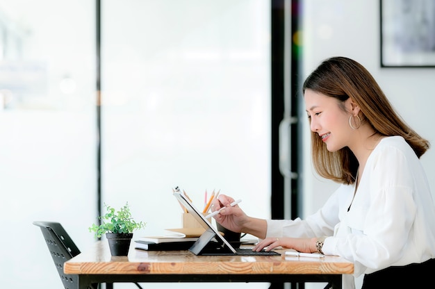 Mulher de negócios nova que usa a escrita digital da caneta na tela da tabuleta ao sentar-se na mesa de escritório no escritório moderno, vista lateral.