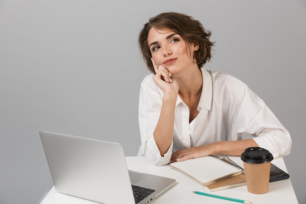 Mulher de negócios posando isolada sobre uma parede cinza, sentada à mesa usando um laptop