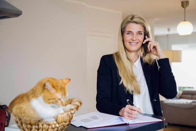 Mulher de negócios respondendo ao telefone celular enquanto o gato está sentado na mesa em casa