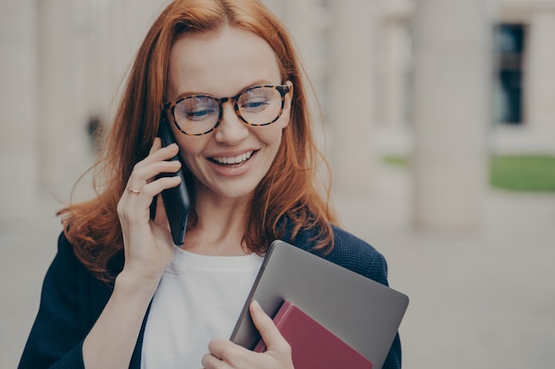 Mulher de negócios ruiva satisfeita com laptop discutindo algo com um parceiro por meio do telefone celular