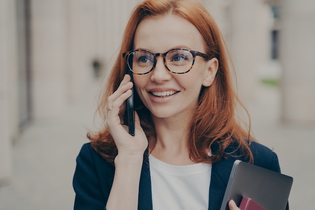 Mulher de negócios ruiva sorridente e positiva usando óculos, ligando para o parceiro, em pé ao ar livre