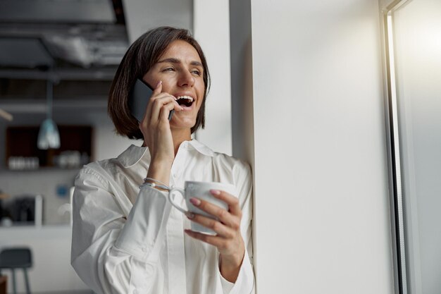 Foto mulher de negócios segurando uma xícara de café e falando ao telefone enquanto está perto da janela no escritório