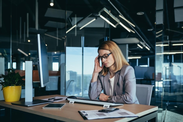 Mulher de negócios sênior tem uma forte dor de cabeça enquanto trabalha em um escritório moderno no computador