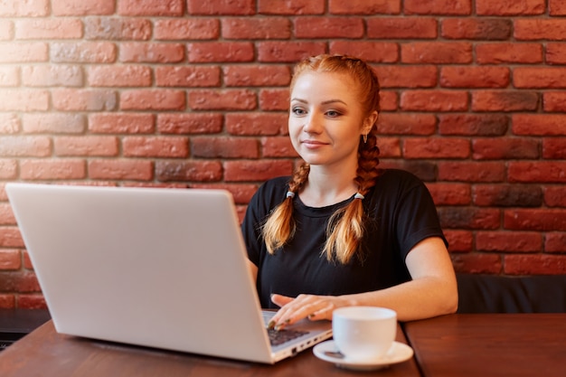 Mulher de negócios sentada contra uma parede de tijolos em um café, trabalhando em seu laptop