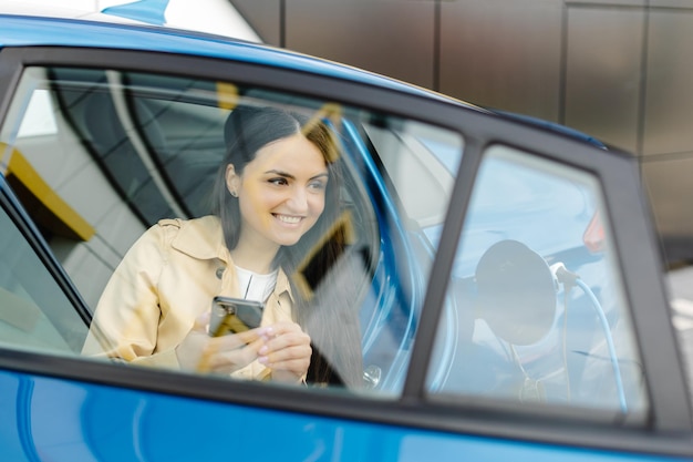 Mulher de negócios sentada dentro de carro elétrico carregando e usando smartphone moderno Tecnologia e conceito de transporte