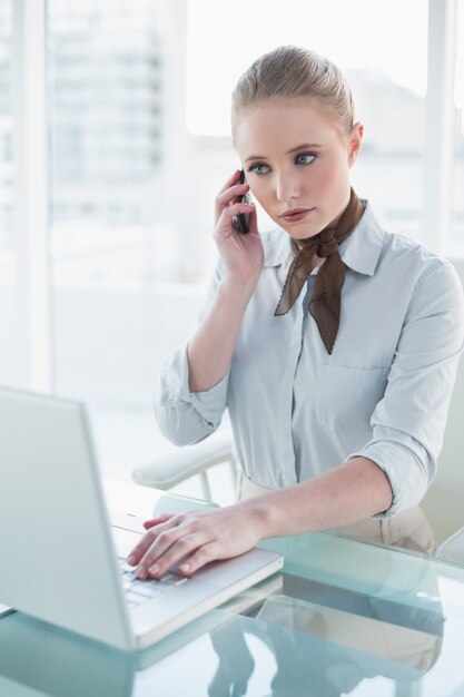 Mulher de negócios séria loira usando laptop e telefonando