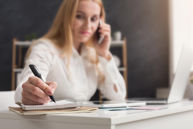 Mulher de negócios sérios no trabalho falando no telefone e fazendo anotações, sentada em seu local de trabalho no escritório, copie o espaço