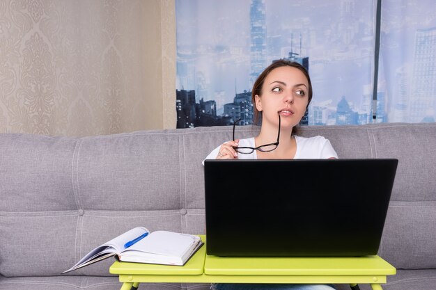 Mulher de negócios sonhadora, sonhando e colocando seus óculos na boca enquanto trabalhava em um laptop, fazendo seus negócios em casa, sentada em um sofá na sala de estar em um ambiente descontraído