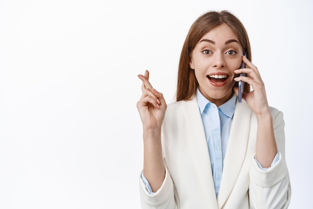 Mulher de negócios sorridente de terno fala no telefone com os dedos cruzados espera notícias de bom presságio falando no smartphone com cara alegre em pé sobre fundo branco