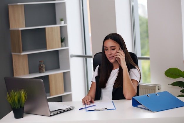 Mulher de negócios sorridente em traje de negócios, sentada na mesa e falando ao telefone no escritório