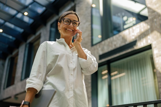 Mulher de negócios sorridente falando ao telefone com um cliente enquanto está em coworking com um laptop