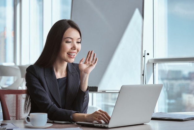 Mulher de negócios sozinha no escritório, sentada à mesa com uma xícara de café quente, tendo uma videochamada no laptop, acenando para a câmera, sorrindo alegre