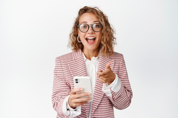 Mulher de negócios surpresa segurando o celular e apontando para a tela, reação a algo incrível no smartphone, fundo branco.