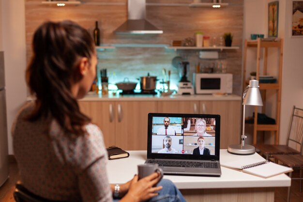 Mulher de negócios tendo uma videoconferência com a equipe durante a meia-noite, usando o laptop na cozinha de casa. Reunião corporativa usando tecnologia moderna, laptop tarde da noite, tecnologia, agência, consultor, trabalho, discussão