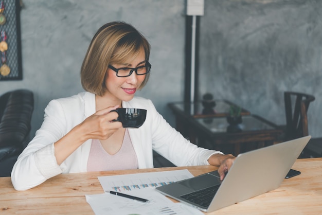 Mulher de negócios tomando café e trabalhando em um laptop, pesquisando na web, navegando em informações, sentada trabalhando na mesa