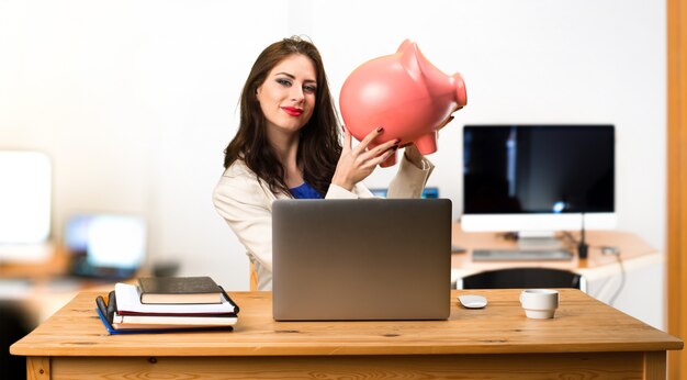 Mulher de negócios, trabalhando com o laptop dela e segurando um piggybank no escritório