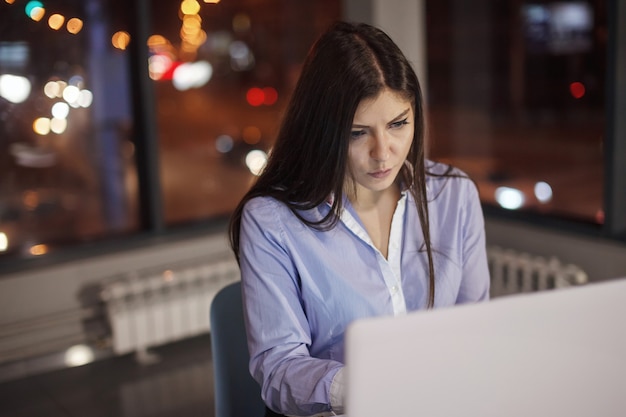 Mulher de negócios trabalhando na mesa com o laptop à noite no escritório. Do lado de fora da janela estão acesas luzes multicoloridas