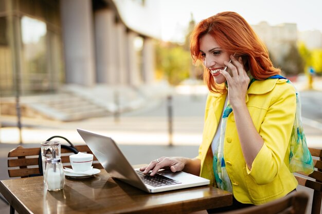 Mulher de negócios trabalhando no laptop e usando telefone inteligente em uma pausa para o café em um café de rua.