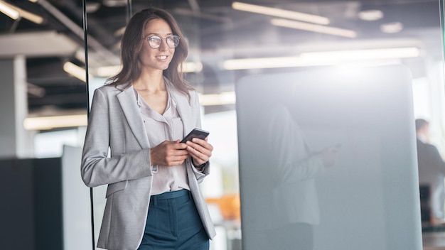 Mulher de negócios usando óculos com celular na mão em pé no escritório moderno