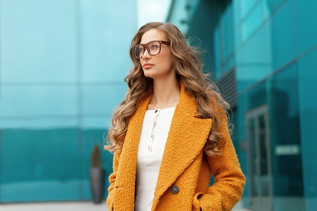 Mulher de negócios vestida de casaco amarelo em pé ao ar livre fundo de edifício corporativo Óculos de pessoa de negócios do sexo feminino caucasiano na rua perto de prédio de escritórios grandes janelas.