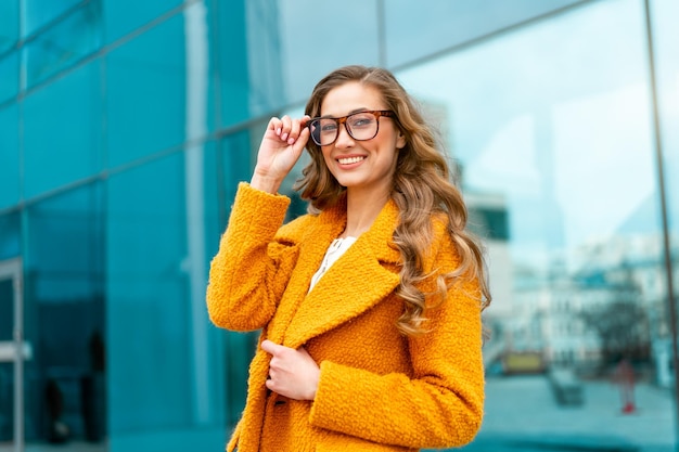 Mulher de negócios vestida de casaco amarelo em pé ao ar livre fundo de edifício corporativo Óculos de pessoa de negócios do sexo feminino caucasiano na rua perto de prédio de escritórios grandes janelas.