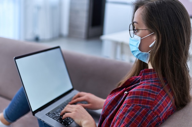 Mulher de óculos e uma máscara de proteção médica enquanto on-line trabalhando em casa no computador durante o auto-isolamento e quarentena.