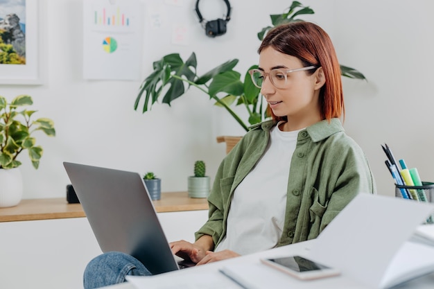 Mulher de óculos trabalhando remotamente em casa com laptop Jovem empresária em um ambiente de trabalho confortável