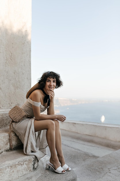 Mulher de olhos castanhos em elegante vestido de verão bege sorri e olha para a câmera morena com saco de palha senta-se nas escadas perto da casa velha com vista para o mar