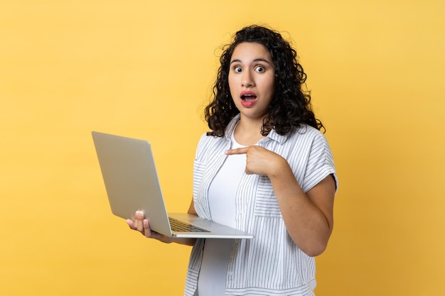 Mulher de pé com o laptop nas mãos e apontando para a tela olhando com olhos grandes e boca aberta