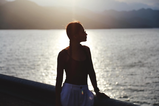 Mulher de pé contra o mar durante o pôr-do-sol