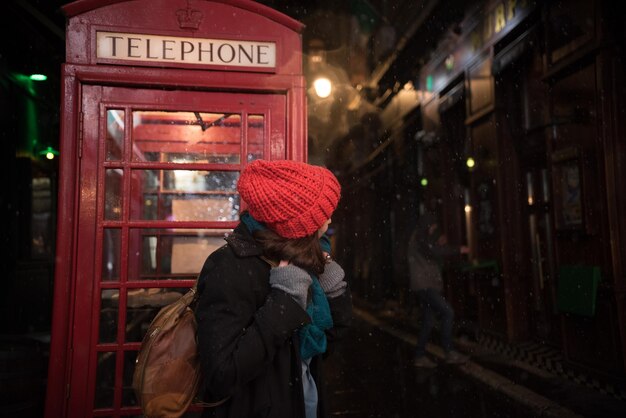 Foto mulher de pé contra uma cabine telefônica à noite