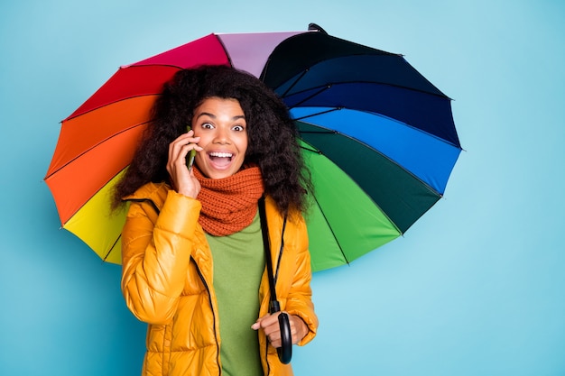 Mulher de pele escura espantada com sombrinha. telefone falando no celular sobre fundo azul