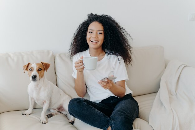 Mulher de pele escura feliz posa em apartamento moderno, senta-se no sofá confortável com animal de estimação, bebe café, usa telefone celular para comunicação on-line, de bom humor, lançando notícias, usa app