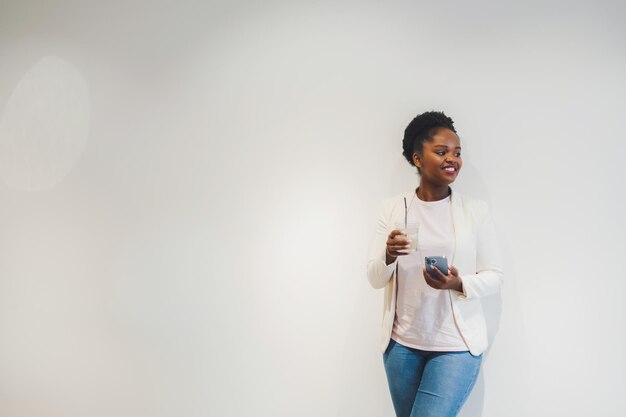 Mulher de pele escura positiva, aproveitando o intervalo do trabalho na cafeteria, bebendo bebidas geladas e mensagens de texto no celular encostado na parede branca com espaço livre para texto copiar espaço