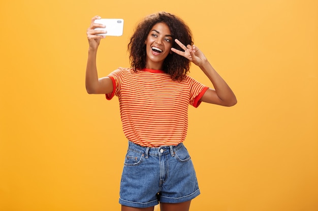 Mulher de pele escura tirando uma selfie e mostrando o sinal da vitória sobre uma parede laranja