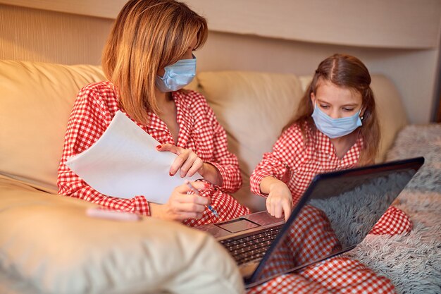 Mulher de pijama com caderno e papéis, trabalhando em casa, usando máscara protetora, enquanto a filha e a filha jogam videogame