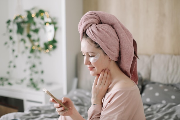 Mulher de pijama com tapa-olhos e toalha na cabeça usando telefone na cama Hábitos matinais e rotina de beleza em casa