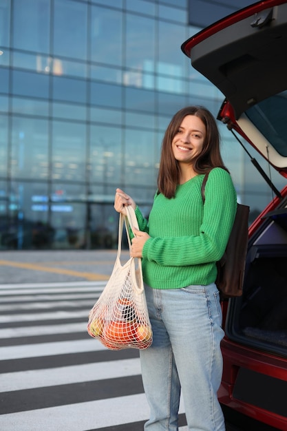 Mulher de pulôver verde segura sacola de compras com mantimentos perto do porta-malas de carros vermelhos