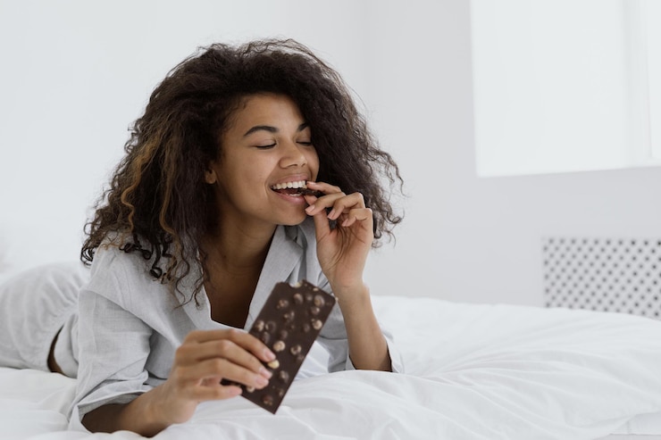 Foto mulher de raça mista a comer chocolate ao leite na cama.