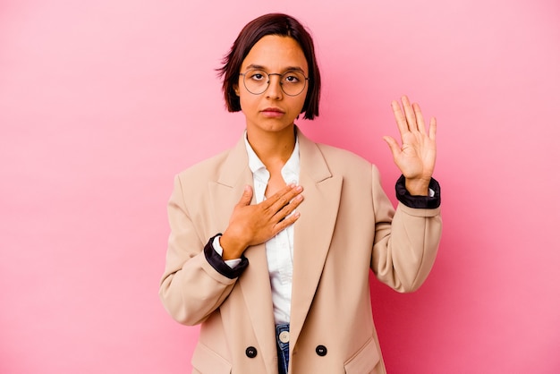 Mulher de raça mista de negócios jovem isolada na parede rosa, fazendo um juramento, colocando a mão no peito.