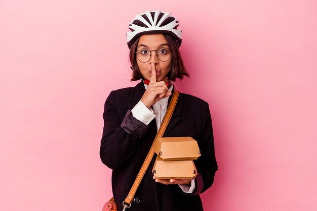Mulher de raça mista de negócios jovens usando capacete de bicicleta segurando hambúrguer isolado no fundo rosa, mantendo um segredo ou pedindo silêncio.