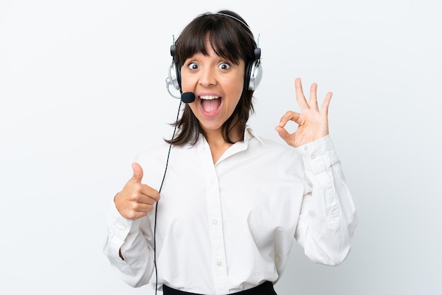 Mulher de raça mista de telemarketing trabalhando com um fone de ouvido isolado no fundo branco, mostrando sinal de ok e polegar para cima gesto