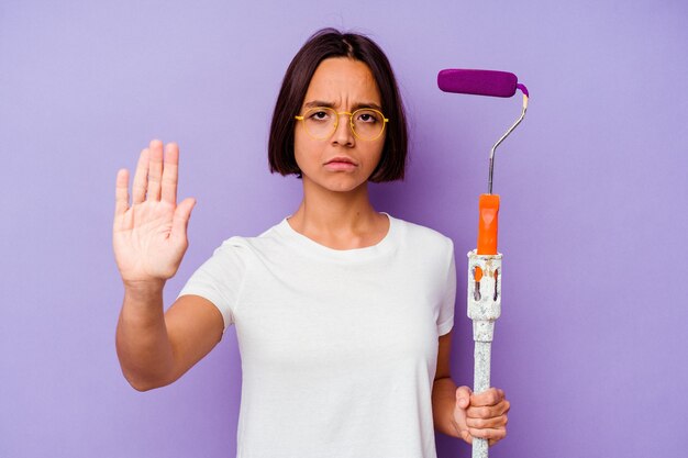 Mulher de raça mista jovem pintor segurando uma vara de tinta isolada no fundo roxo em pé com a mão estendida, mostrando o sinal de pare, impedindo-o.