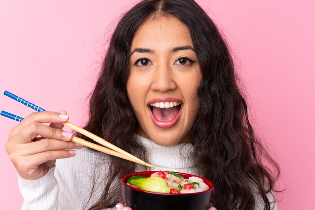 Mulher de raça mista, segurando uma tigela cheia de macarrão sobre parede rosa isolada