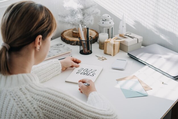 Mulher de resolução de ano novo de metas em suéter branco escrevendo metas de texto no bloco de notas aberto sobre a mesa